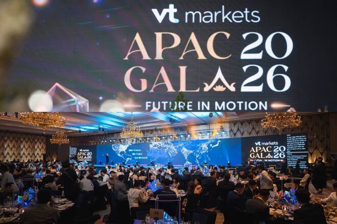 A large, elegant gala event with round banquet tables filled with formally dressed attendees. A stage at the front features a wide digital screen displaying “VT Markets APAC Gala 2026 – Future in Motion,” with blue and gold lighting, chandeliers overhead, and a world map graphic in the background. The atmosphere is upscale and celebratory, with people dining and watching the presentation.