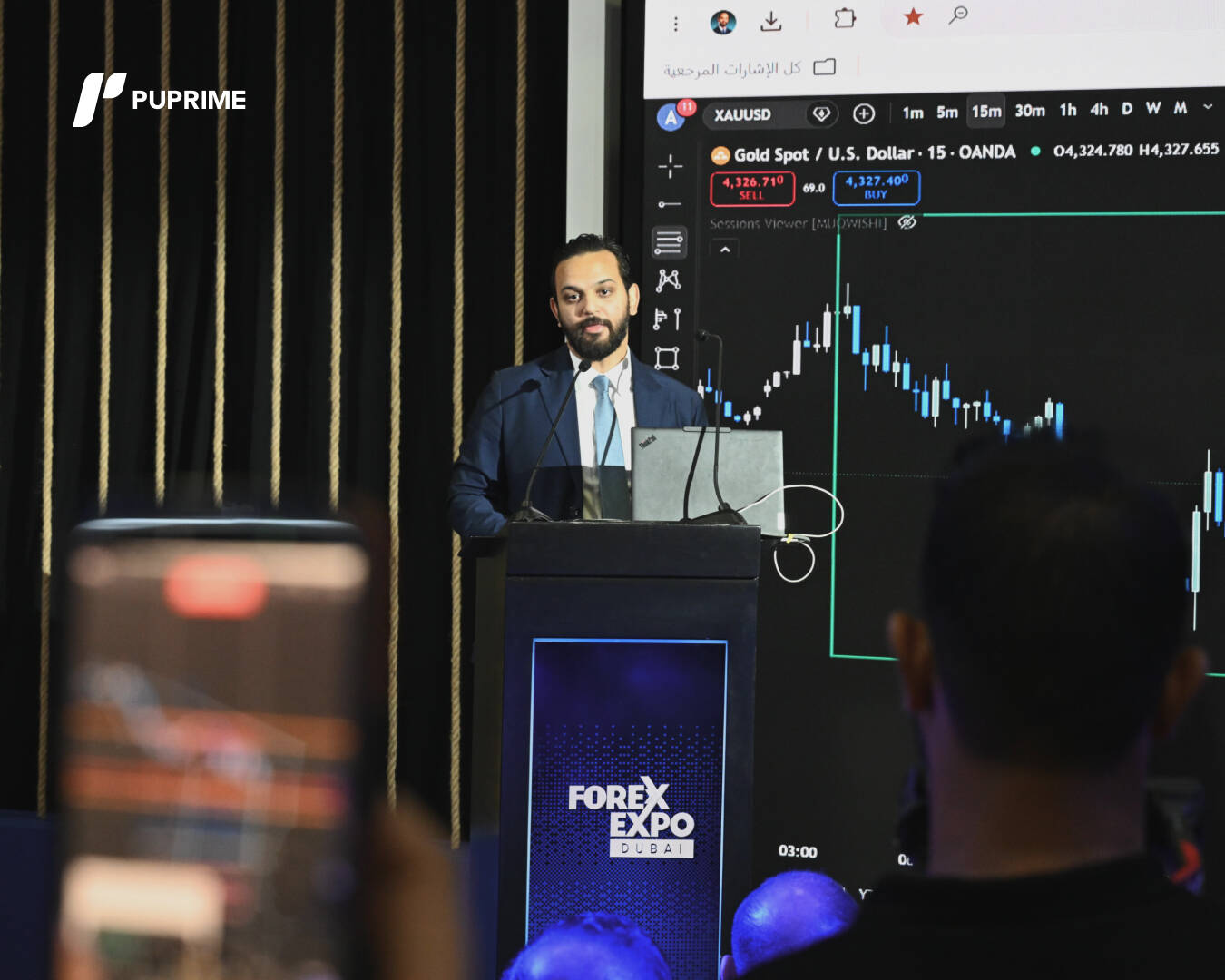 A PU Prime speaker delivering a presentation at the Forex Expo Dubai podium in front of a live Gold/USD candlestick trading chart.