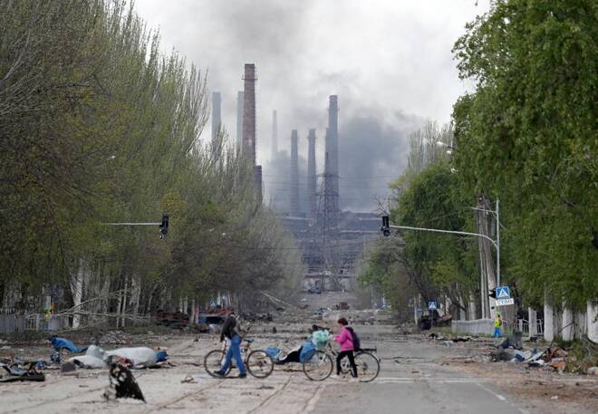 Varias personas cruzan con sus bicicletas una calle de la ciudad ucraniana de Mariúpol, mientras salen columnas de humo del complejo metalúrgico Azovstal al fondo.